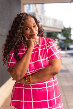 Person in pink and white checkered dress poses expressively against outdoor backdrop.
