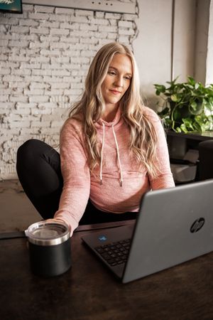 Person in pink sweater works on laptop against white brick wall with plants.