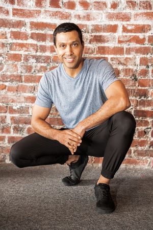 Man in gray t-shirt and black pants posing against brick wall in casual fitness photoshoot.