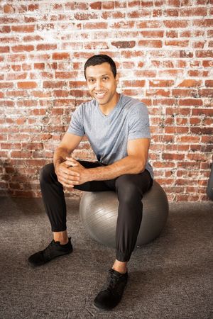 Person sitting on exercise ball against rustic brick wall wearing gray shirt and black pants.