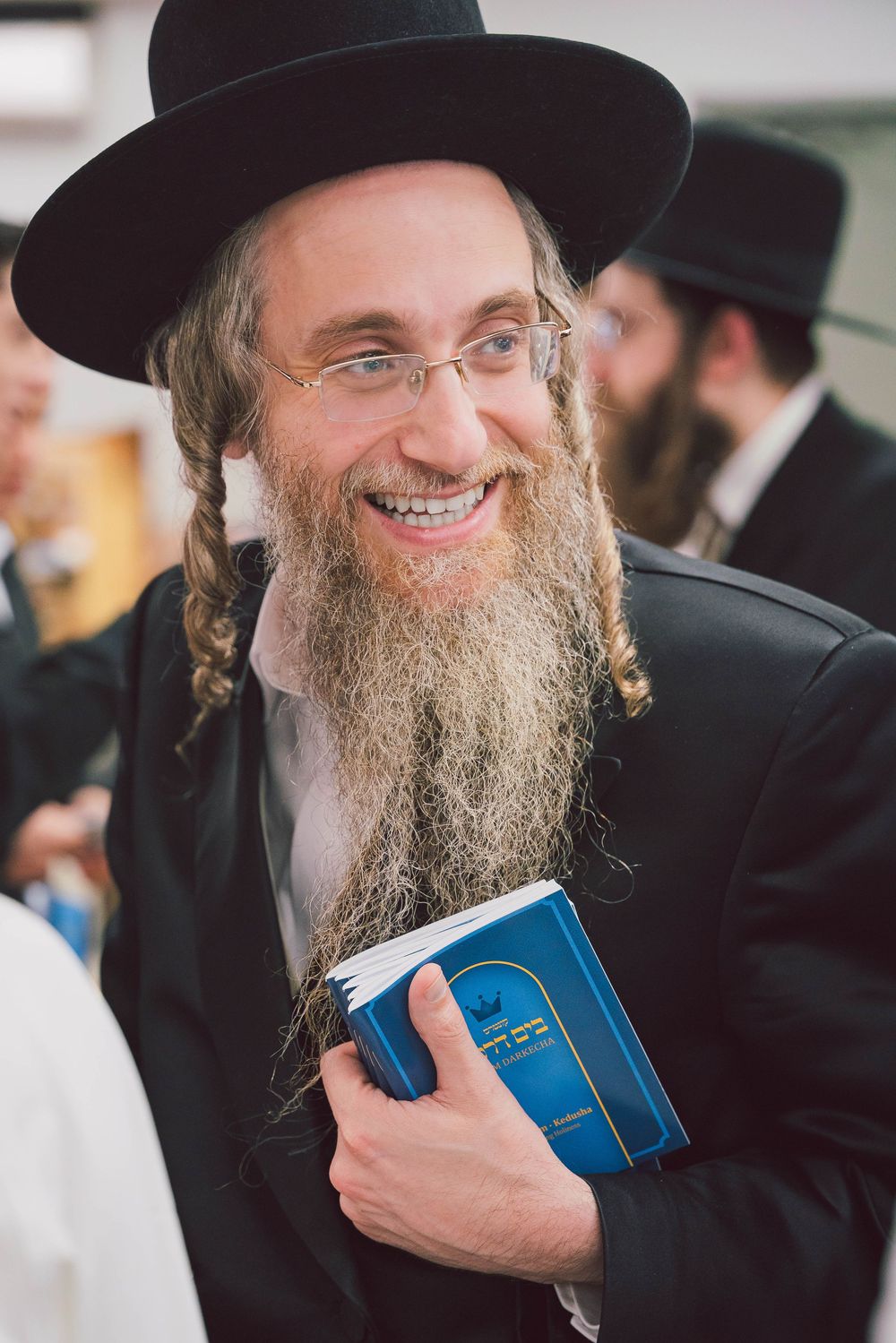 A smiling Orthodox Jewish man in traditional black attire and beard holds a blue book close to his chest.