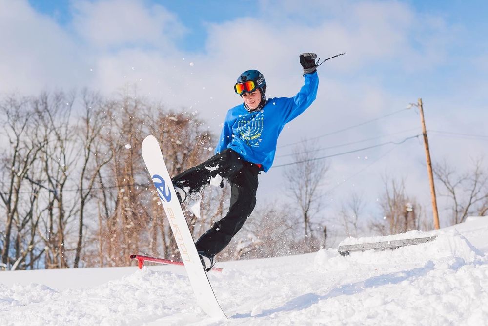 A snowboarder in a blue jacket performs a trick jump at a snowy ski resort with bare trees in the background.