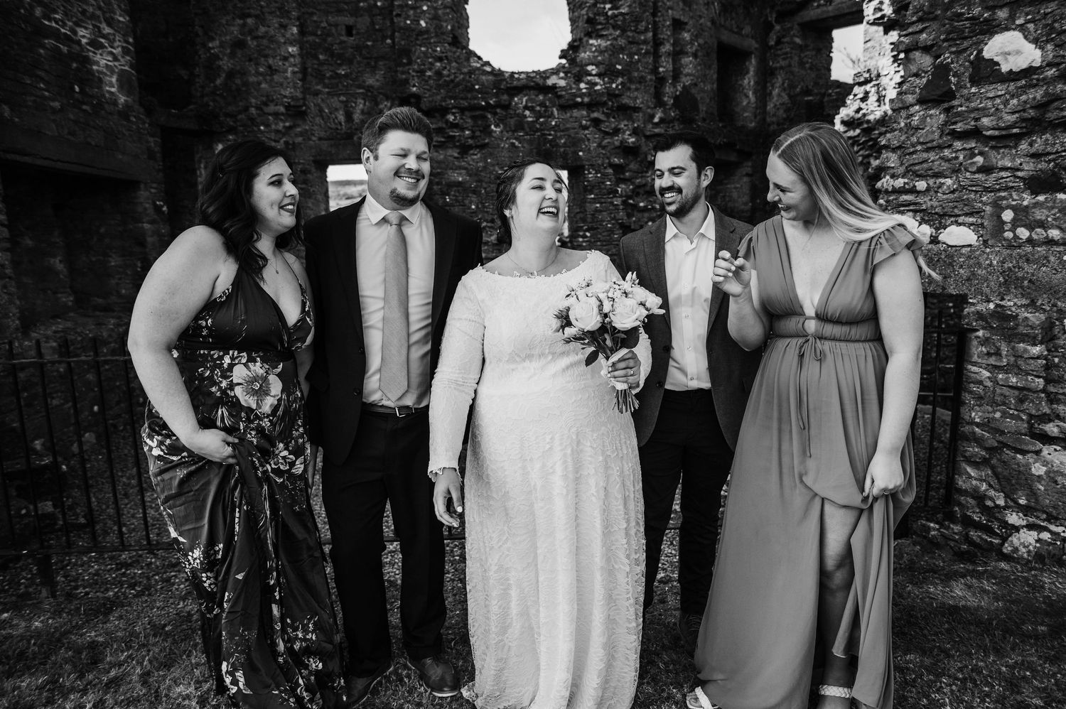 Wedding party in formal attire laughs together while standing against a rustic stone wall outdoors.