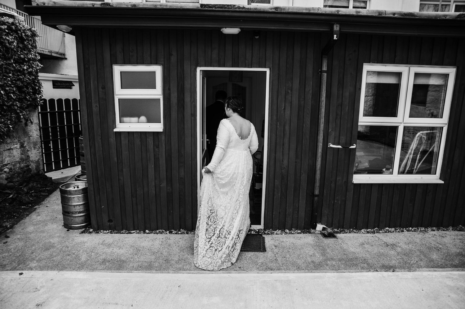 A figure in a white lace dress enters a dark wooden outbuilding with white-trimmed windows.