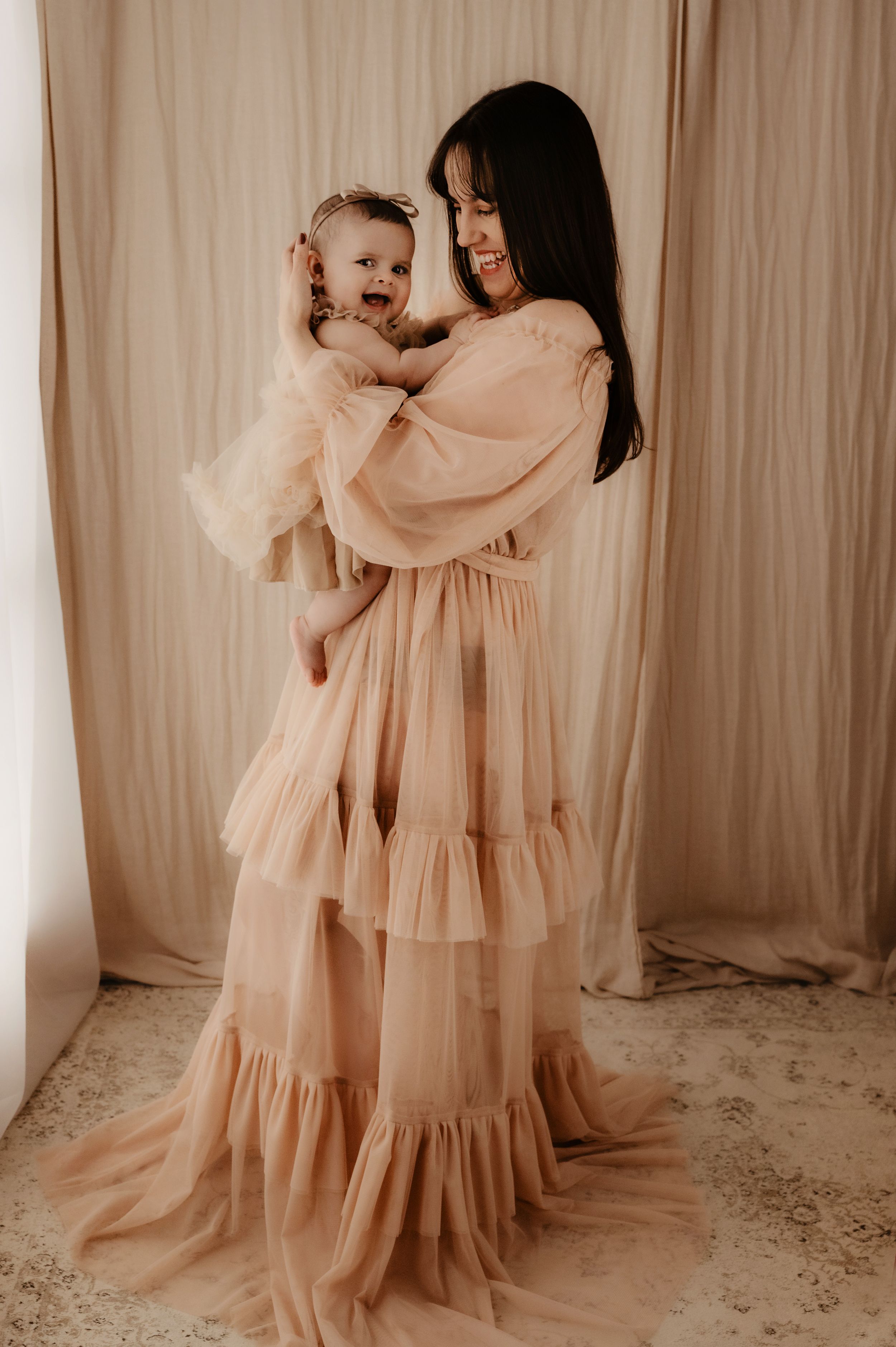 Mother and baby in matching flowy peach dresses pose against neutral curtain backdrop.