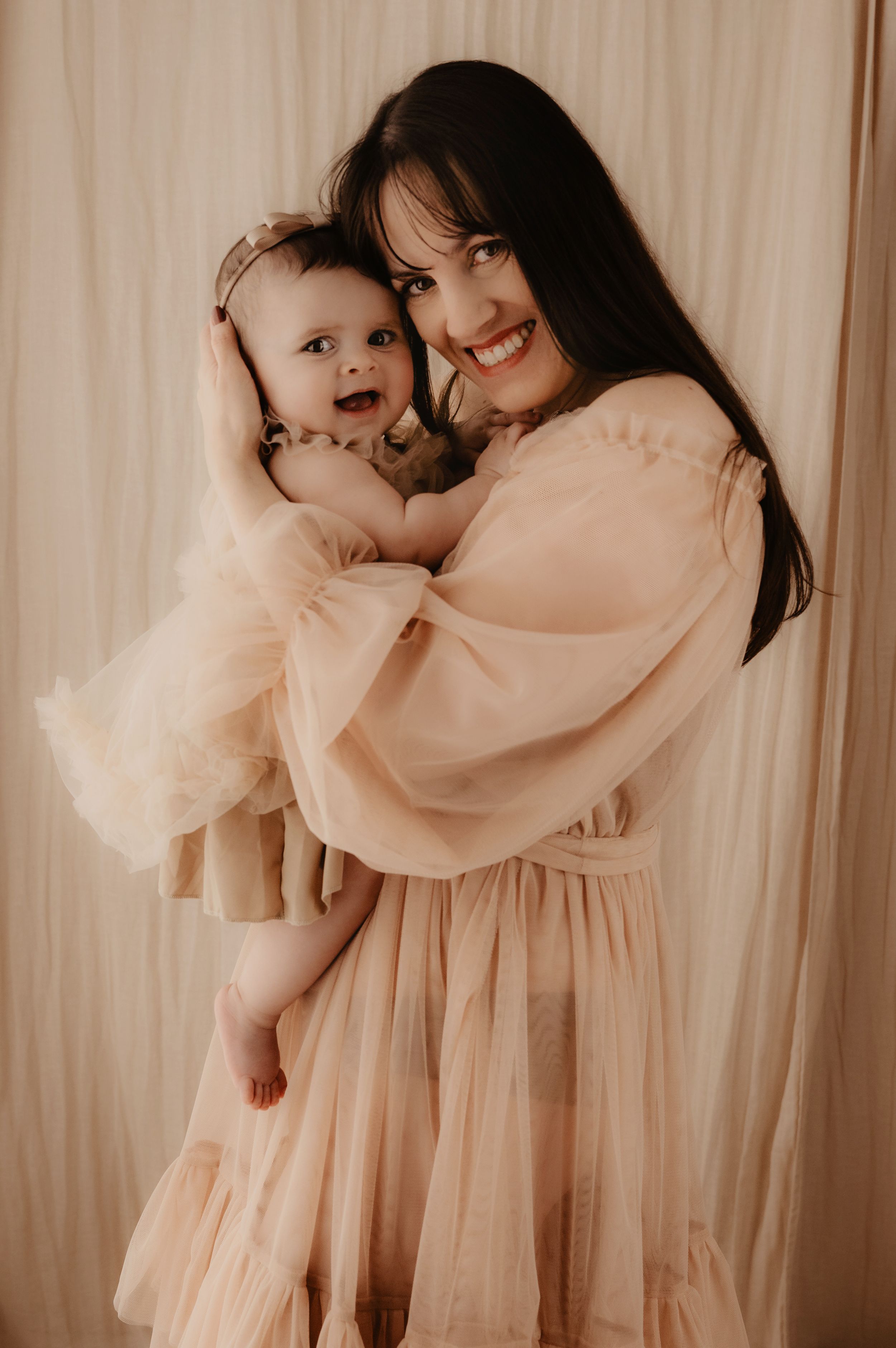 Mother and child in peach-colored dresses share tender embraces against a curtained backdrop.
