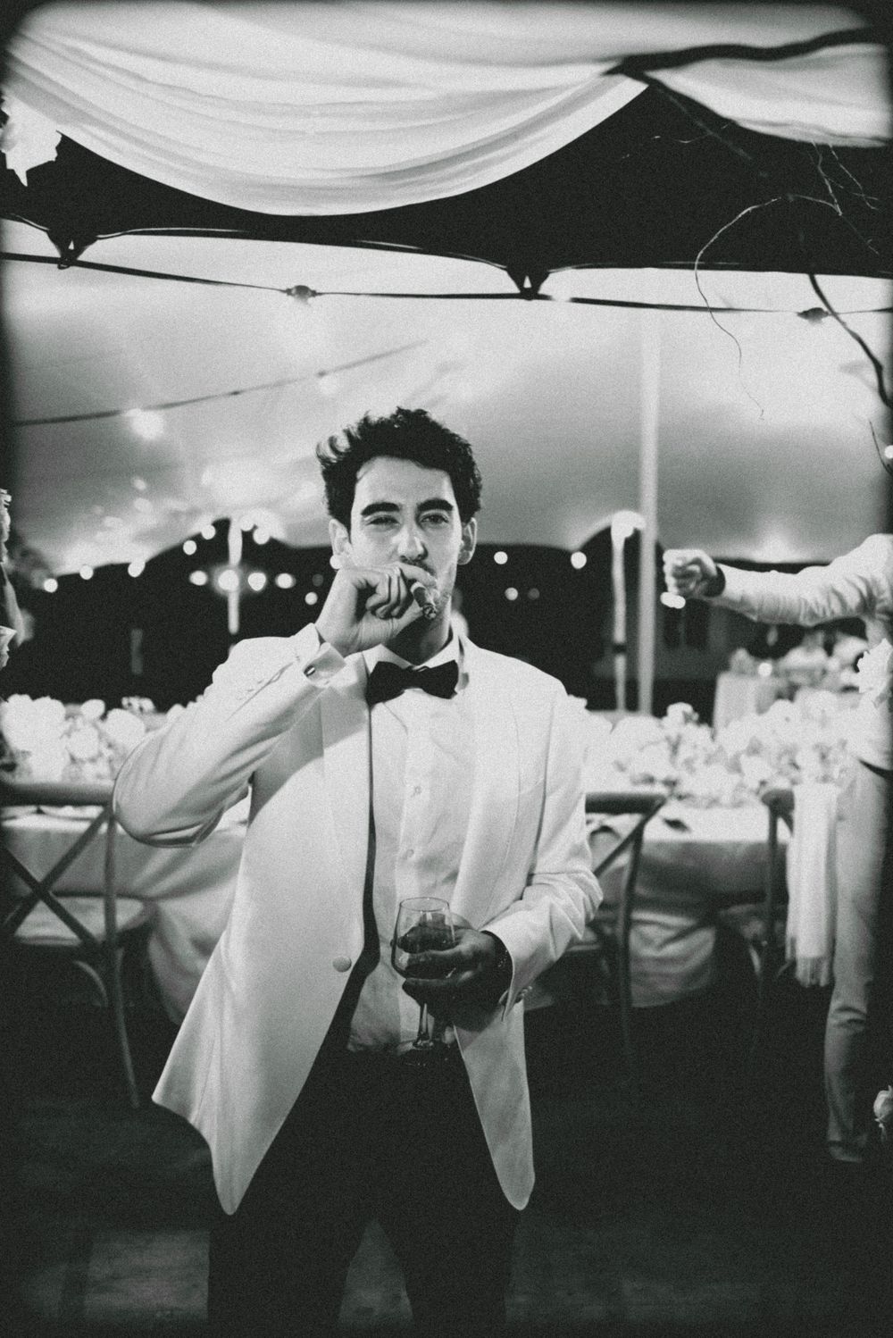 A black and white photo of someone in a white tuxedo jacket smoking a cigarette at an elegant event under draped fabric.
