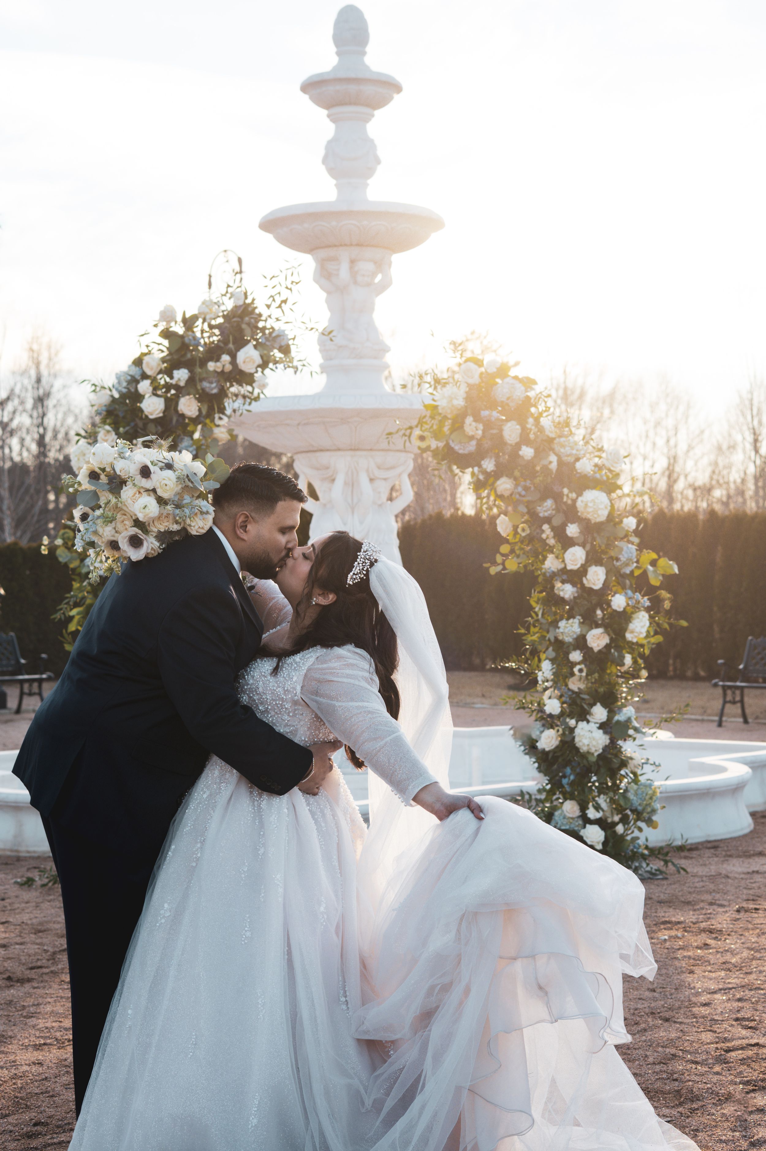 A romantic wedding moment at sunset with a white fountain and floral arch decoration in the background.