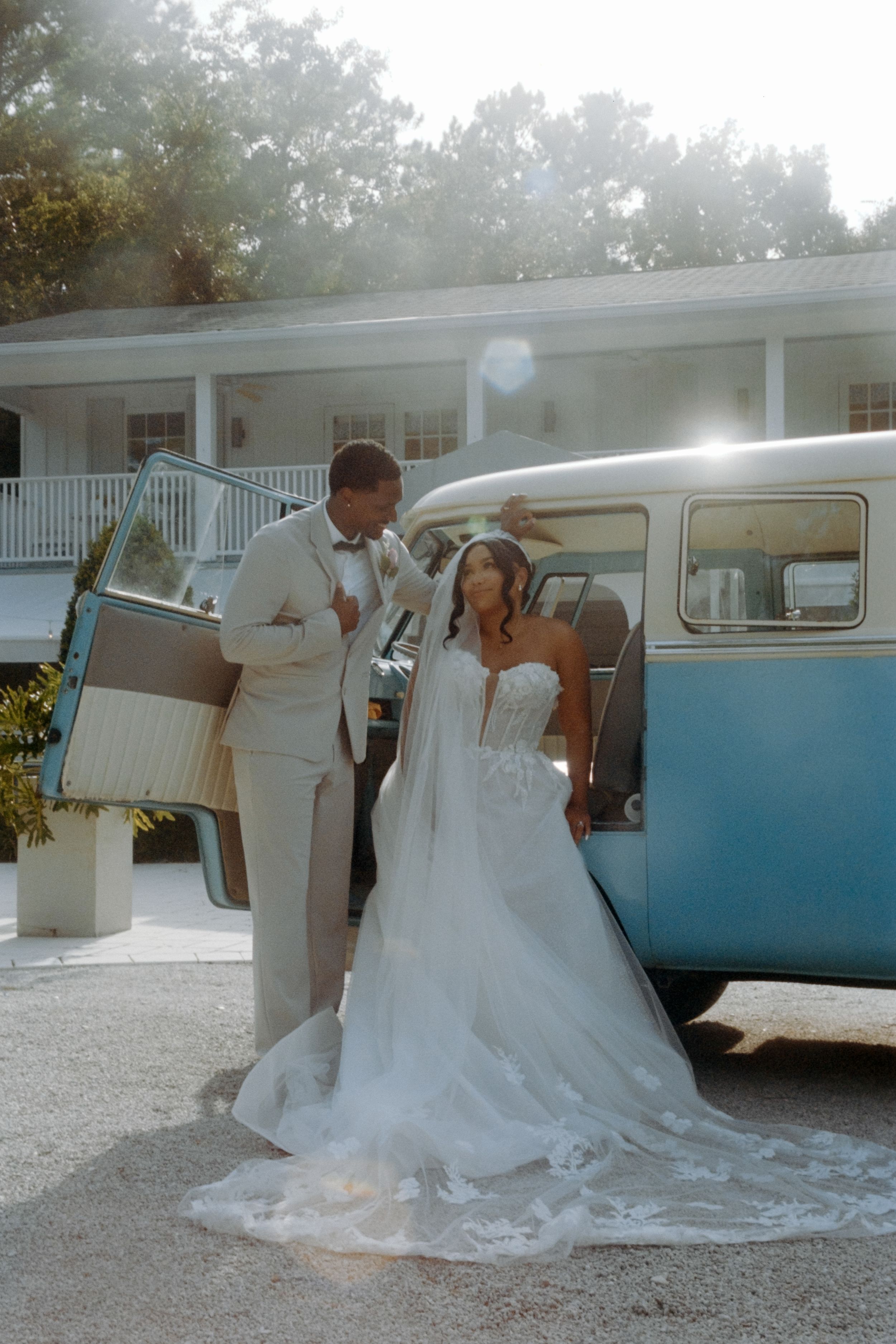 Wedding celebration with bride and groom sharing a romantic moment next to a vintage blue and white VW van in sunlit setting.