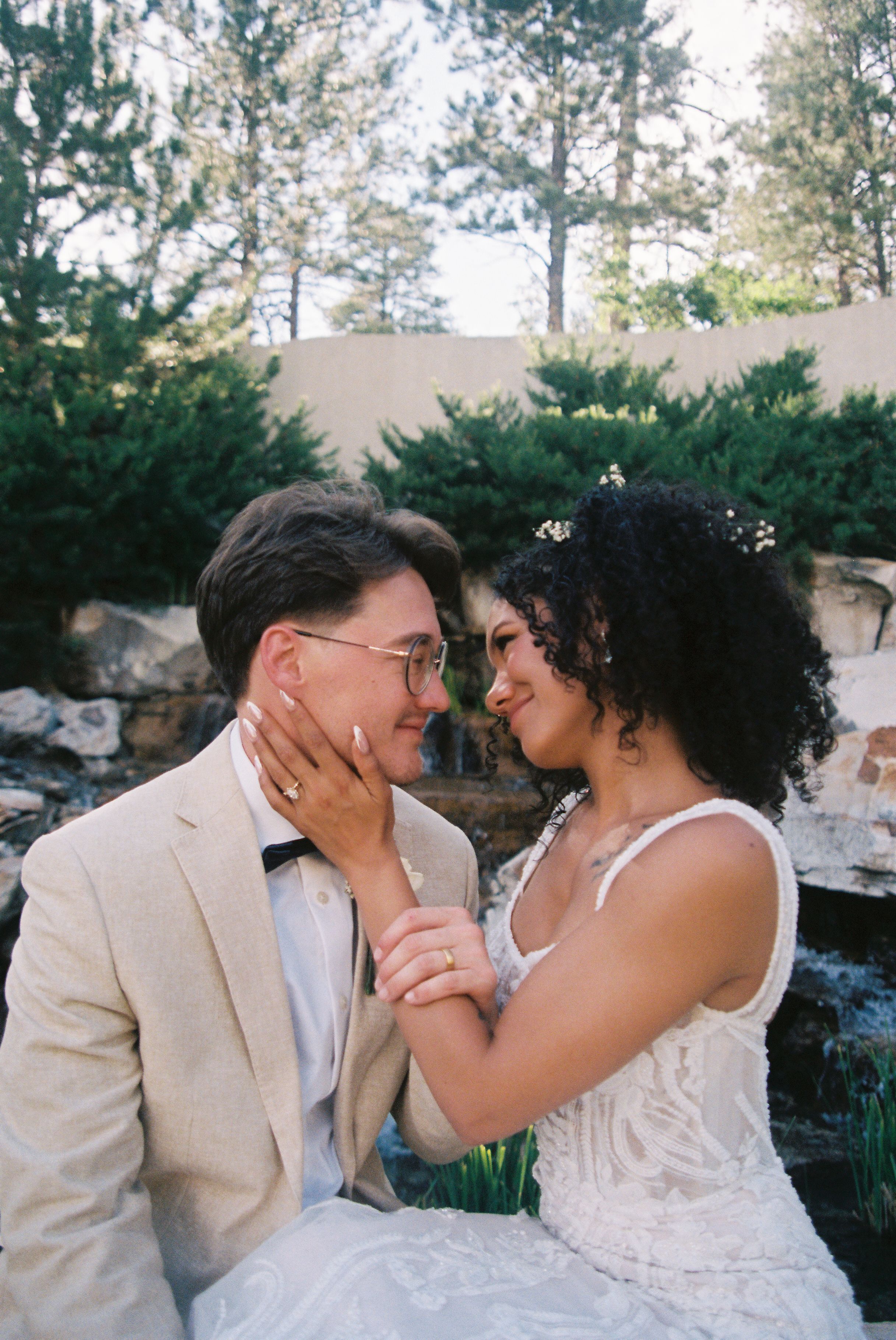 A romantic outdoor wedding moment between a couple in cream attire surrounded by evergreen trees and stone landscaping.