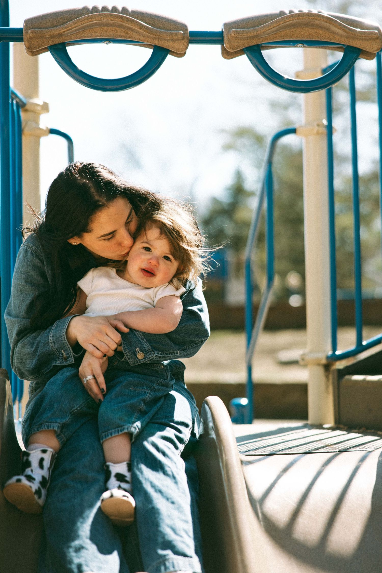 Mother kissing toddler on playground slide, both wearing denim jackets, sunny day at park.