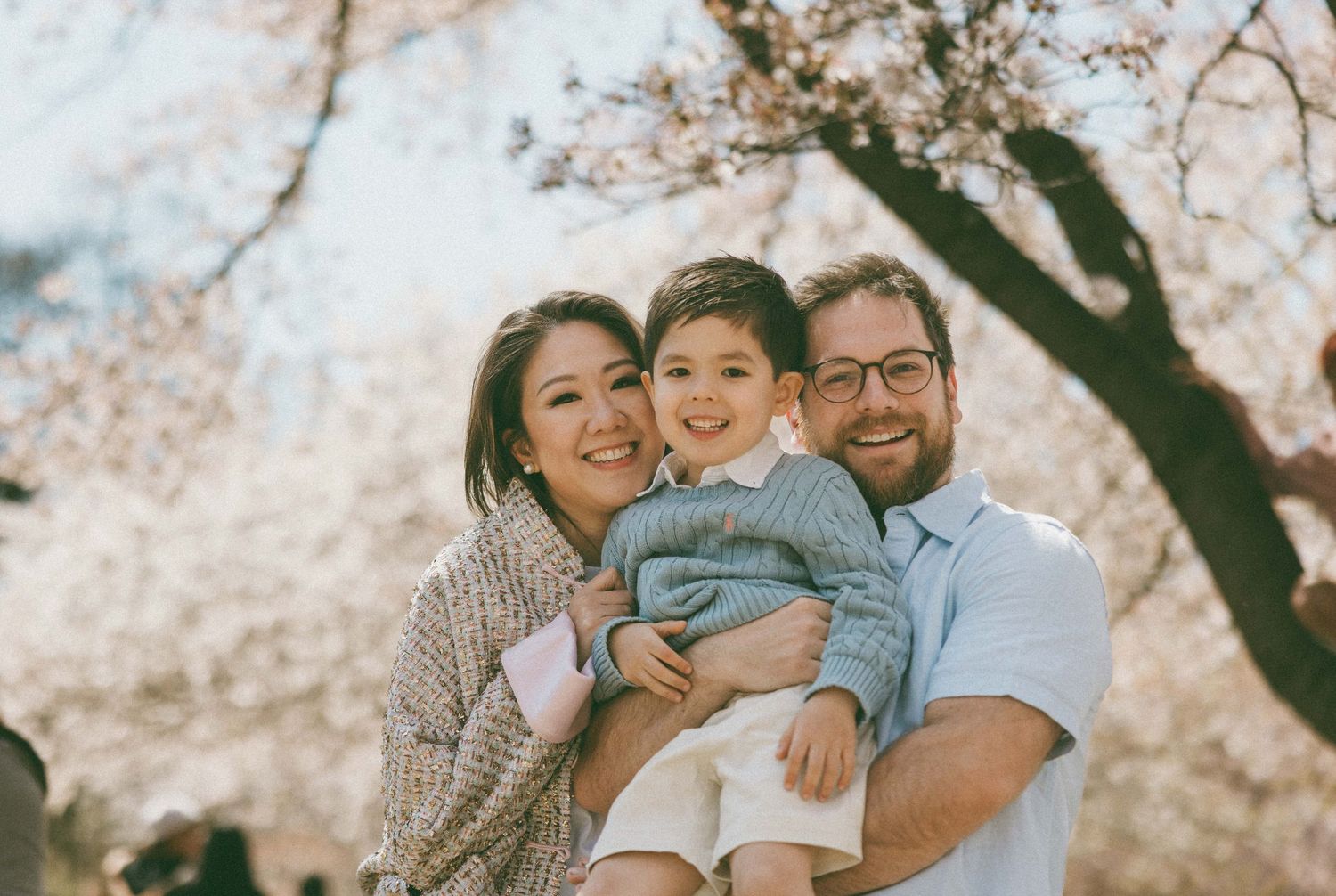 A happy family of three poses among blooming cherry blossom trees, parents holding their young child outdoors.