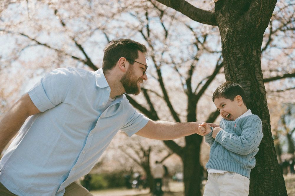 A father and young son share a playful moment under blooming cherry blossom trees in a park.