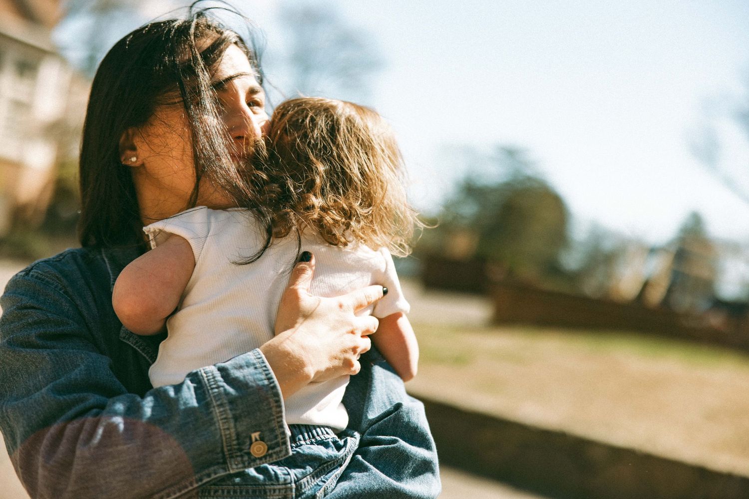 A mother tenderly kisses her curly-haired toddler while holding them outdoors in warm sunlight.