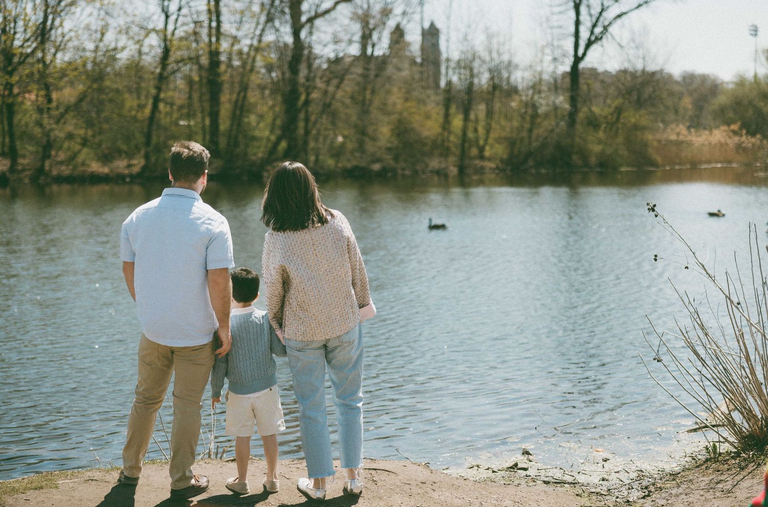 Family of three holding hands while looking at a peaceful lake surrounded by trees.