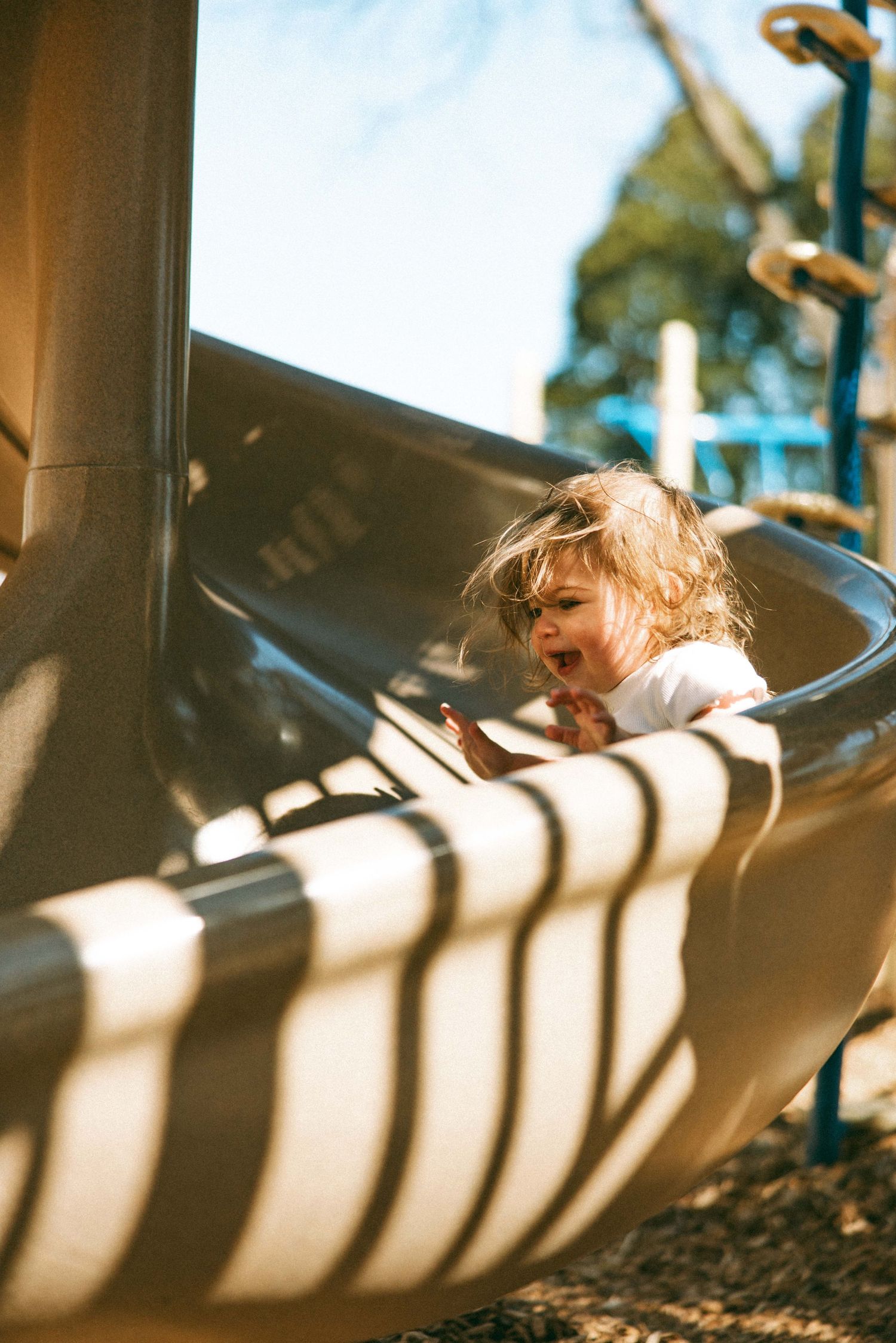 Toddler with curly hair laughing while sliding down a playground slide on a sunny day.