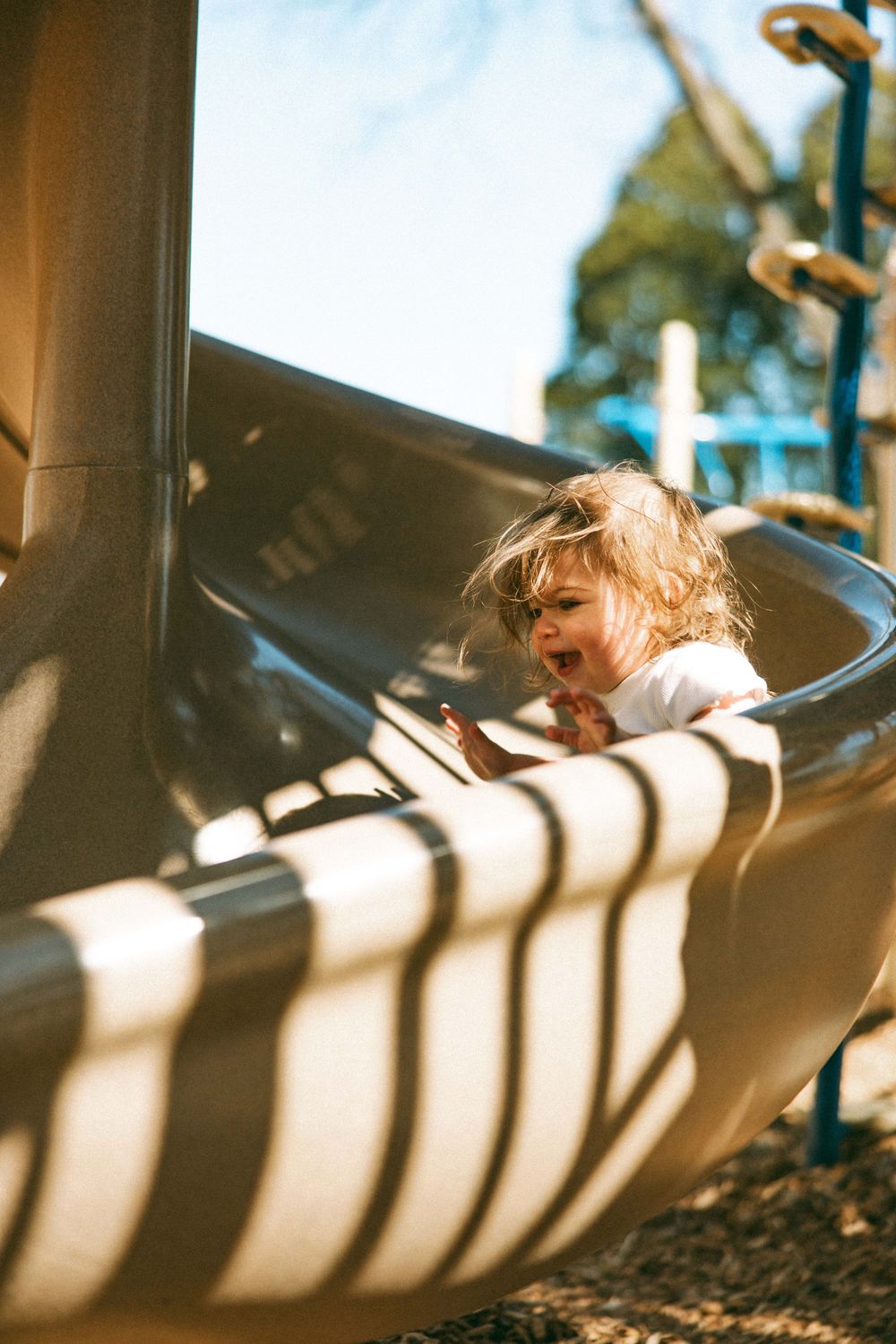Toddler with curly hair laughing while sliding down a playground slide on a sunny day.
