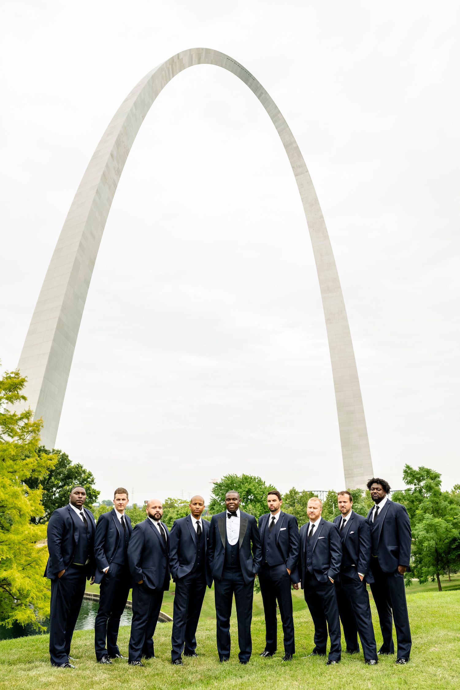 Wedding party members in navy suits pose in front of the iconic St. Louis Gateway Arch on a sunny day.