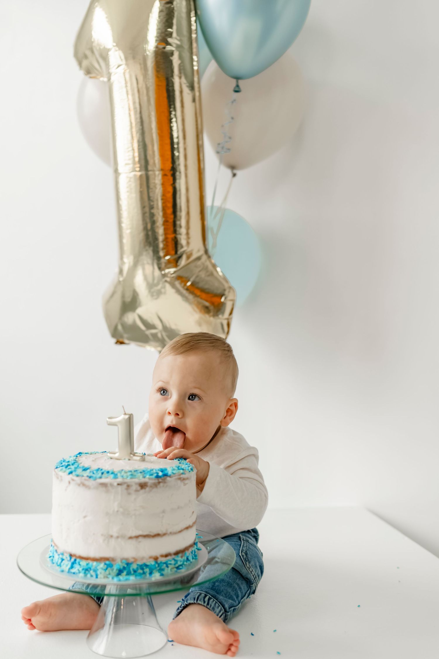 Baby sits next to first birthday cake with blue frosting and silver number one balloon.