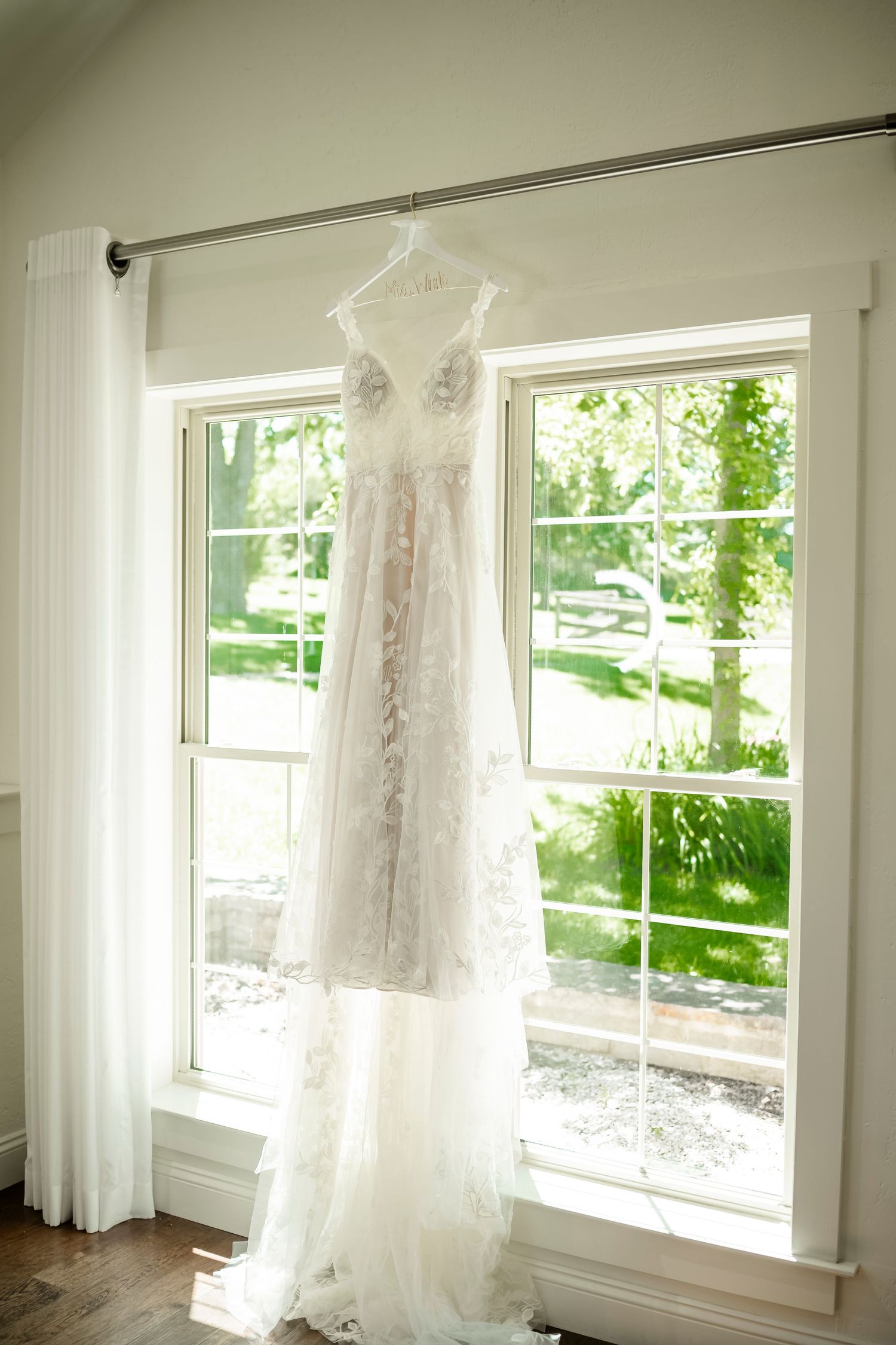 White wedding dress hangs in bright window with natural light streaming through sheer curtains.