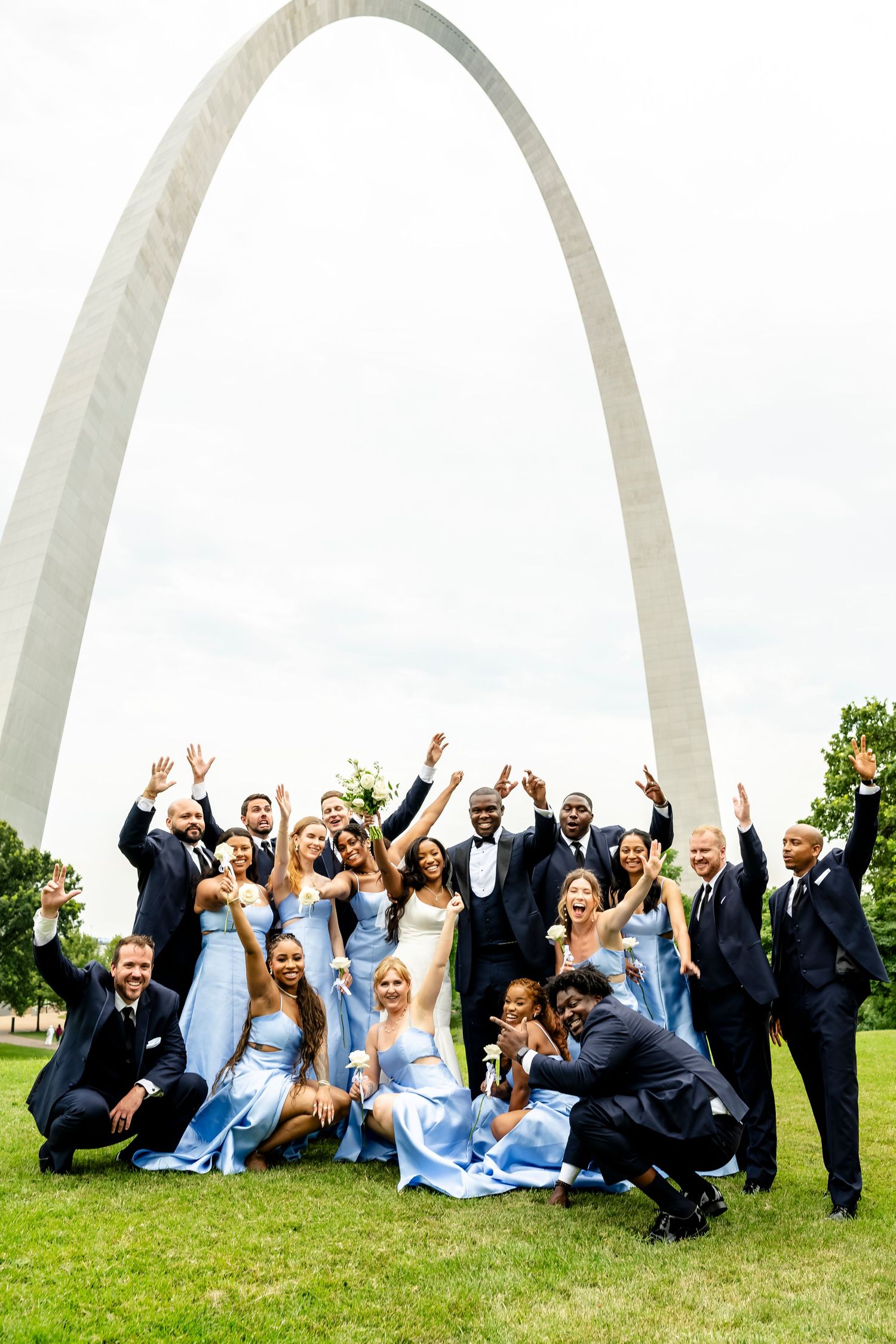 Wedding party celebrates joyfully on grass with the Gateway Arch in background wearing blue dresses and dark suits.