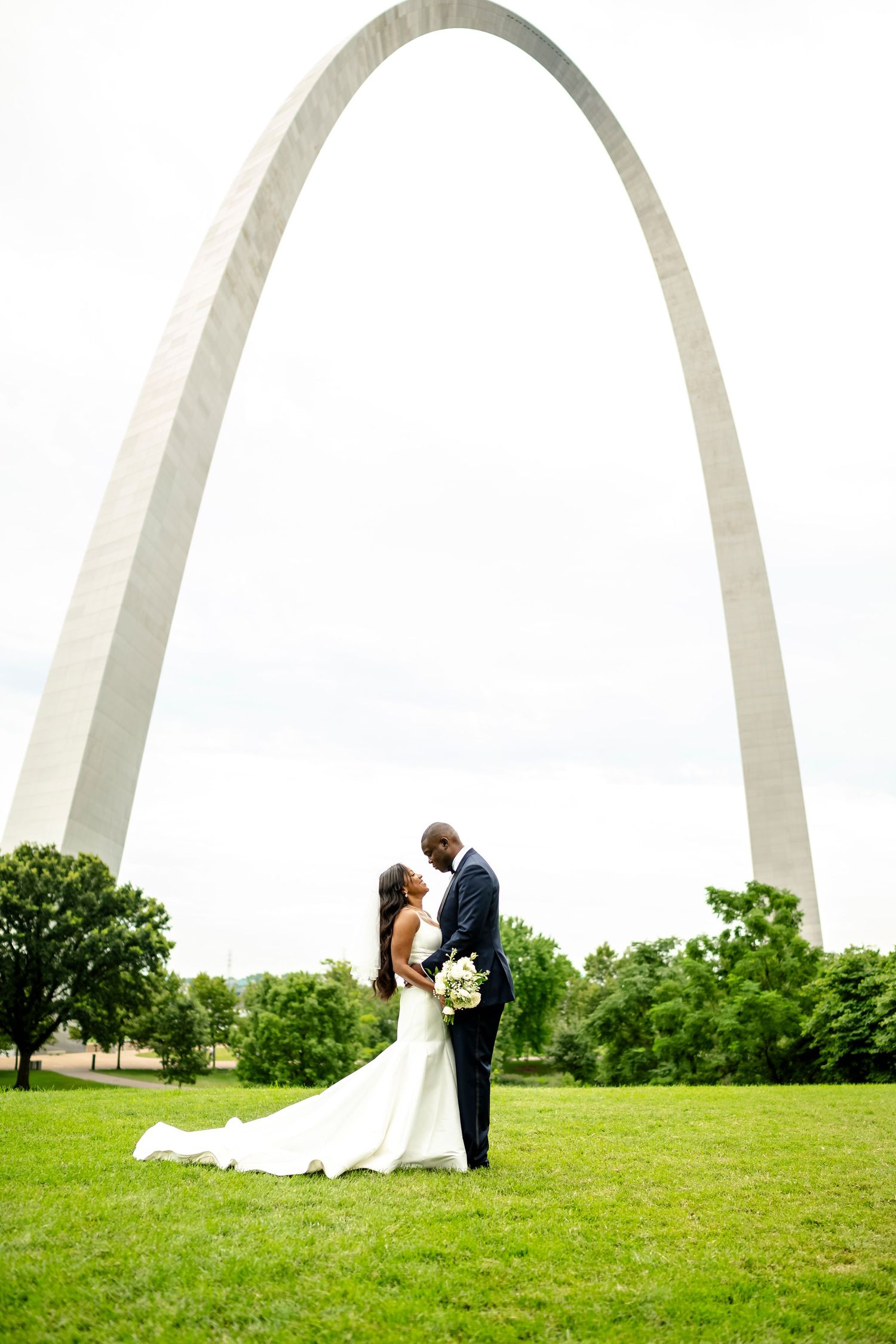 Romantic wedding couple poses on grassy hill with iconic St. Louis Arch creating dramatic backdrop.