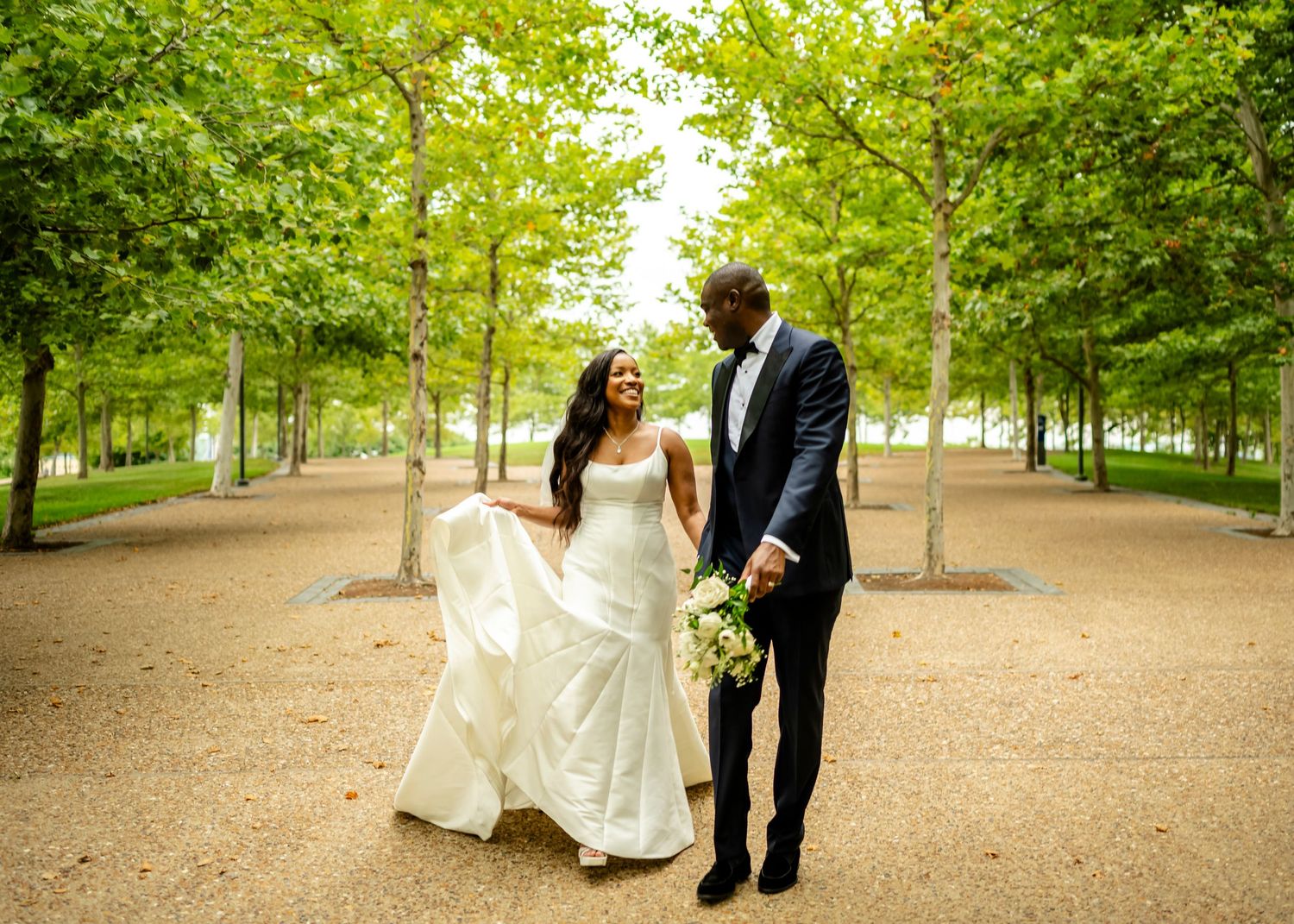 Wedding couple walks through tree-lined path in elegant attire during outdoor photo session.