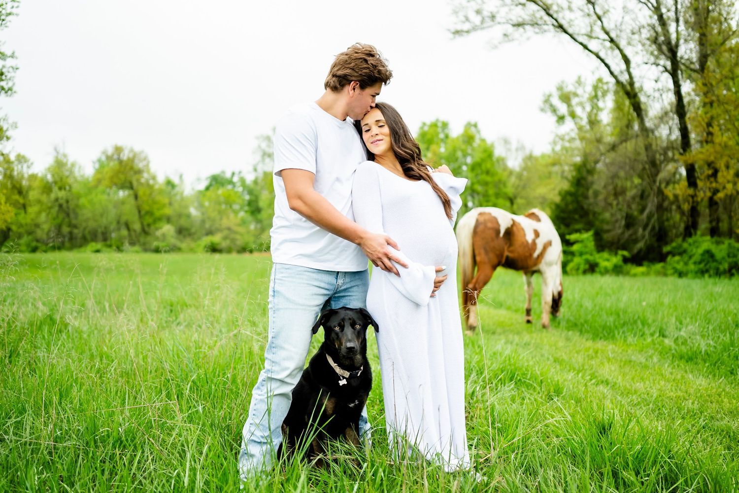 Couple poses in meadow with black dog and horse grazing in background.