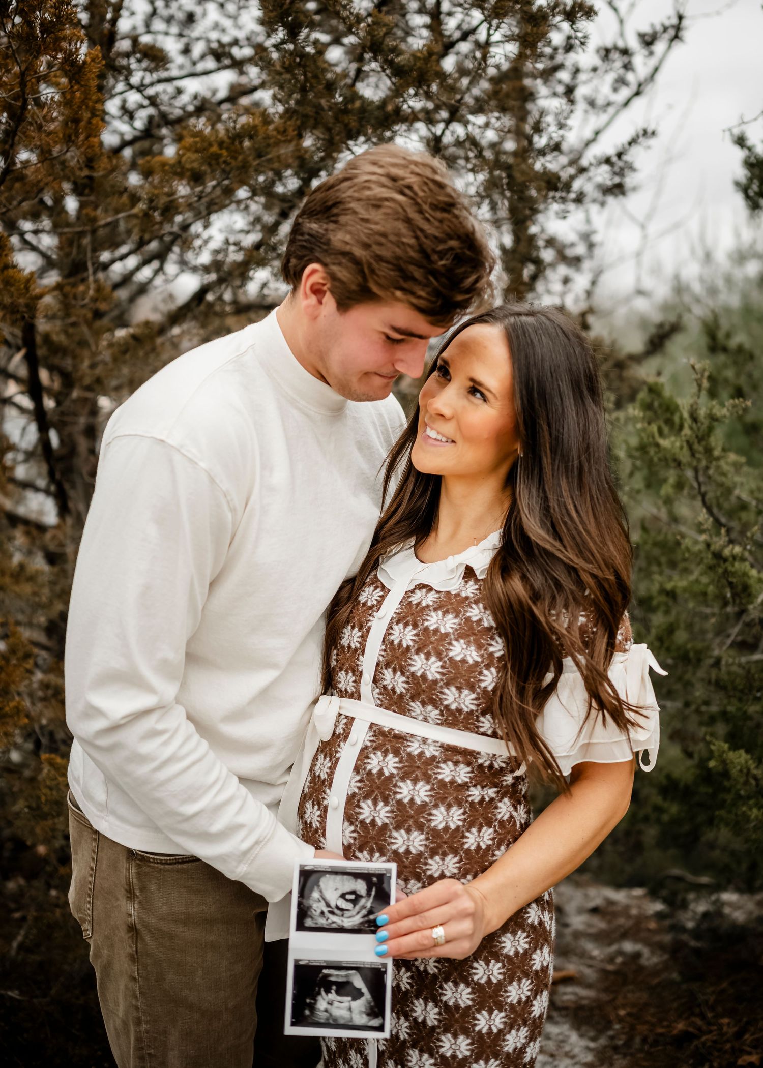 Couple poses with ultrasound photos in outdoor autumn setting.