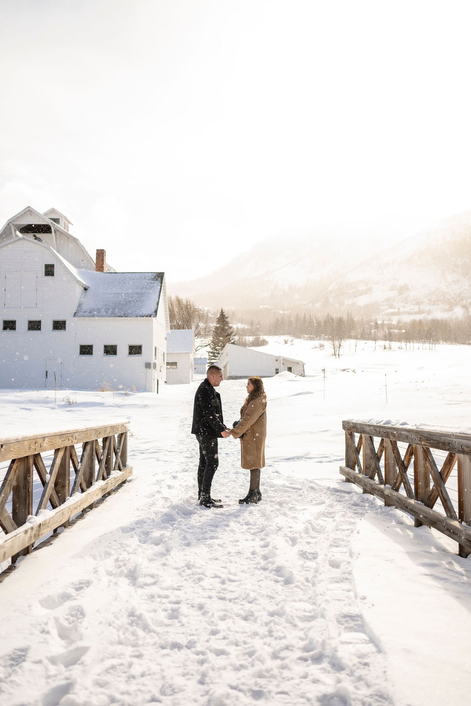 A couple stands on a snowy wooden bridge with a white barn and mountains in the background.