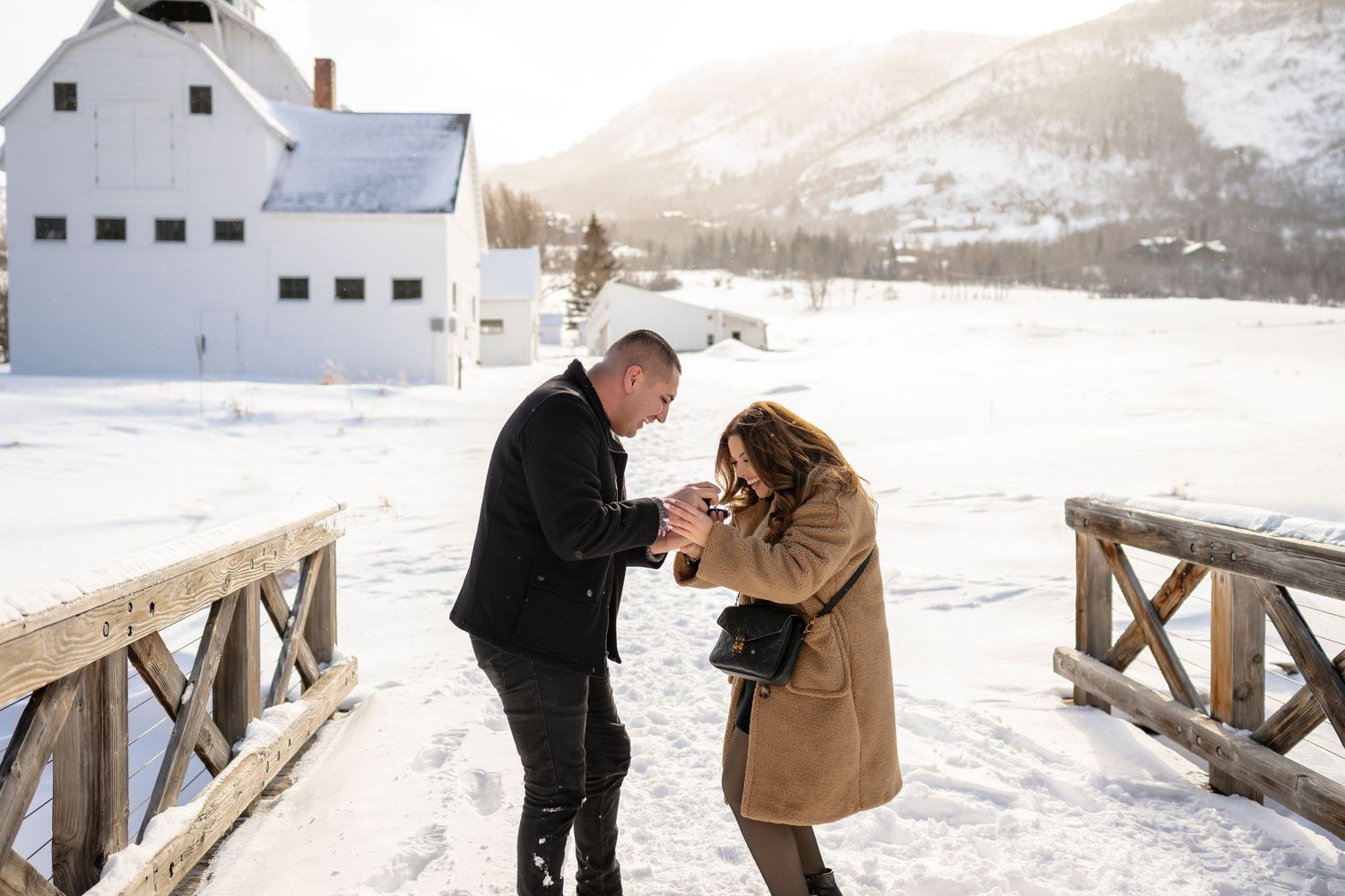 A romantic winter proposal moment on a snow-covered bridge with a white barn in the distance.