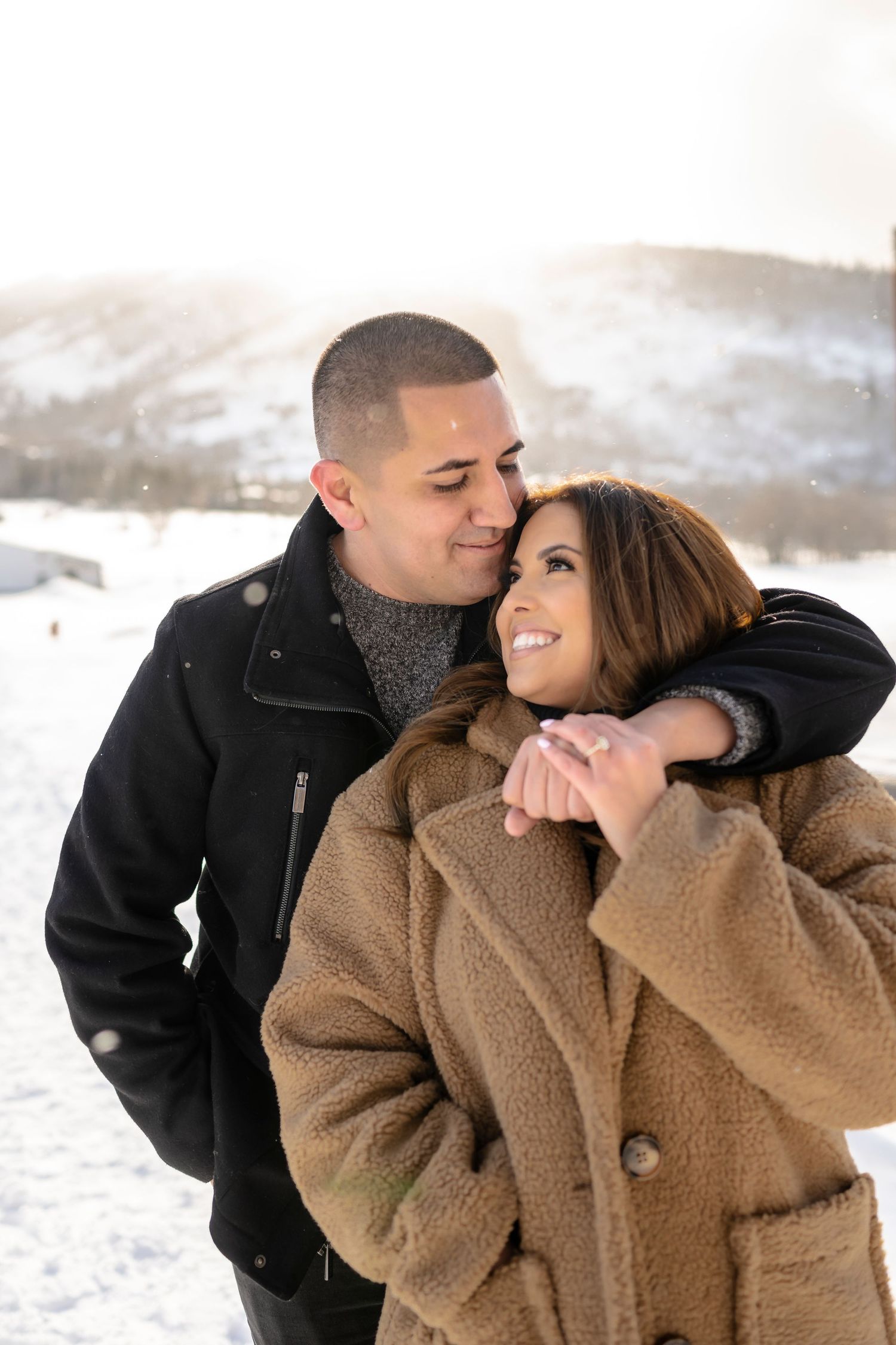 A couple embraces in a cozy winter setting with snow-covered mountains behind them.
