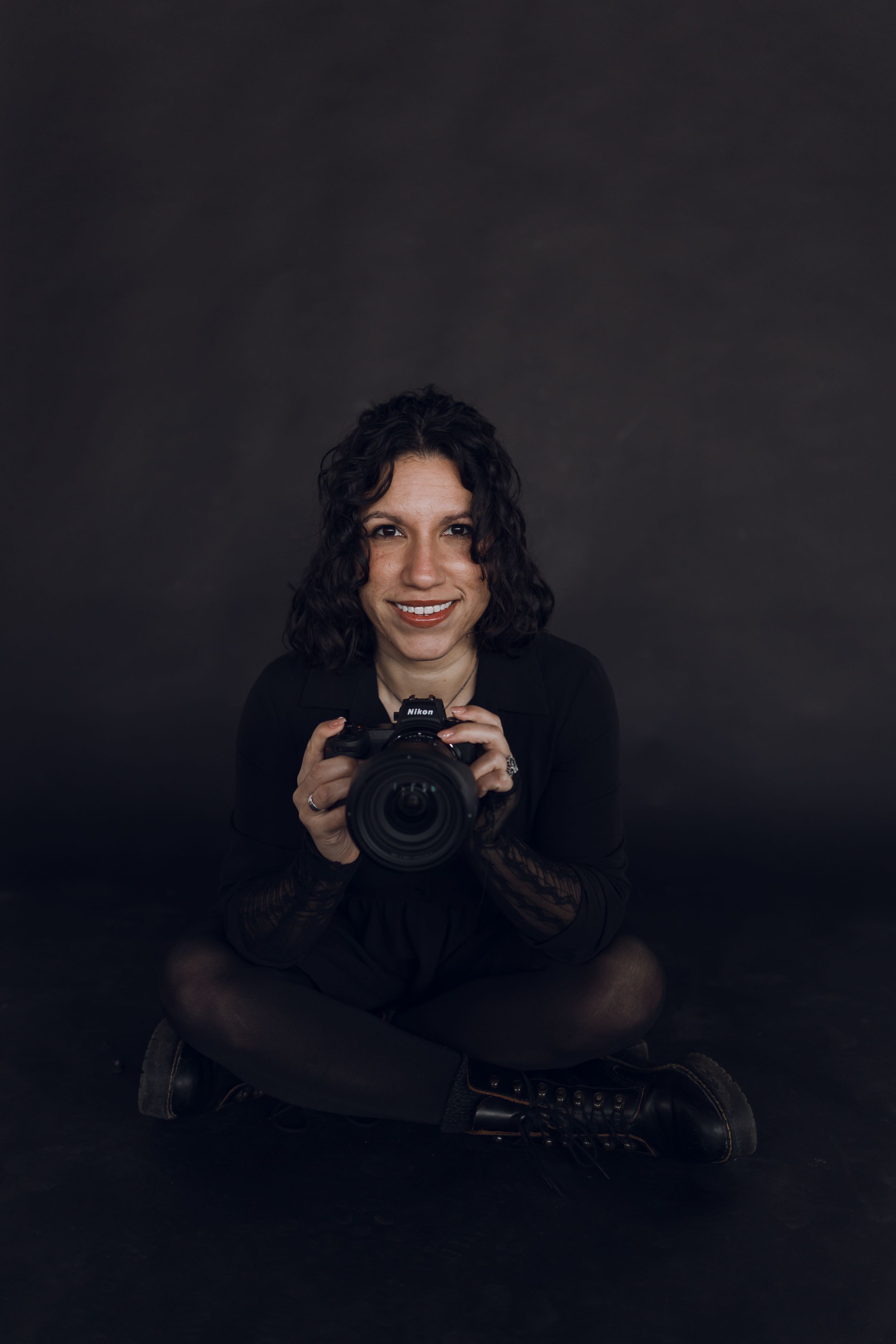 Professional photographer in all black holding a DSLR camera smiles enthusiastically on a tree-lined sidewalk by brick building.