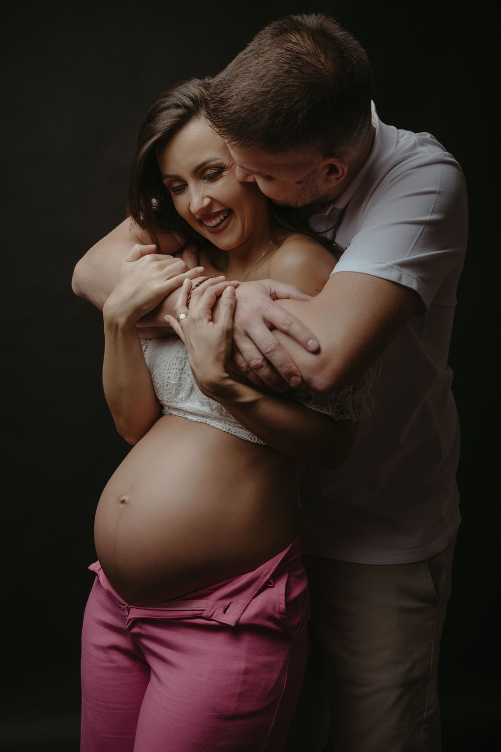 A couple shares a tender moment as a man embraces a smiling pregnant woman in pink pants against a dark background.