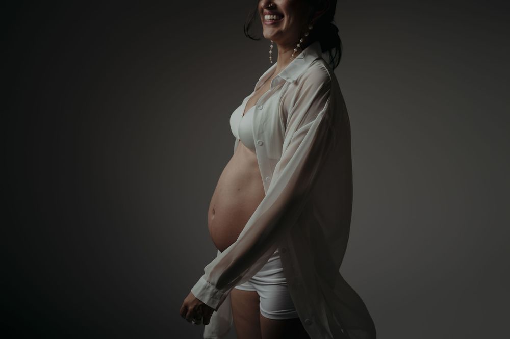 Smiling pregnant woman in white sheer top and shorts showcasing baby bump in a studio maternity photo.