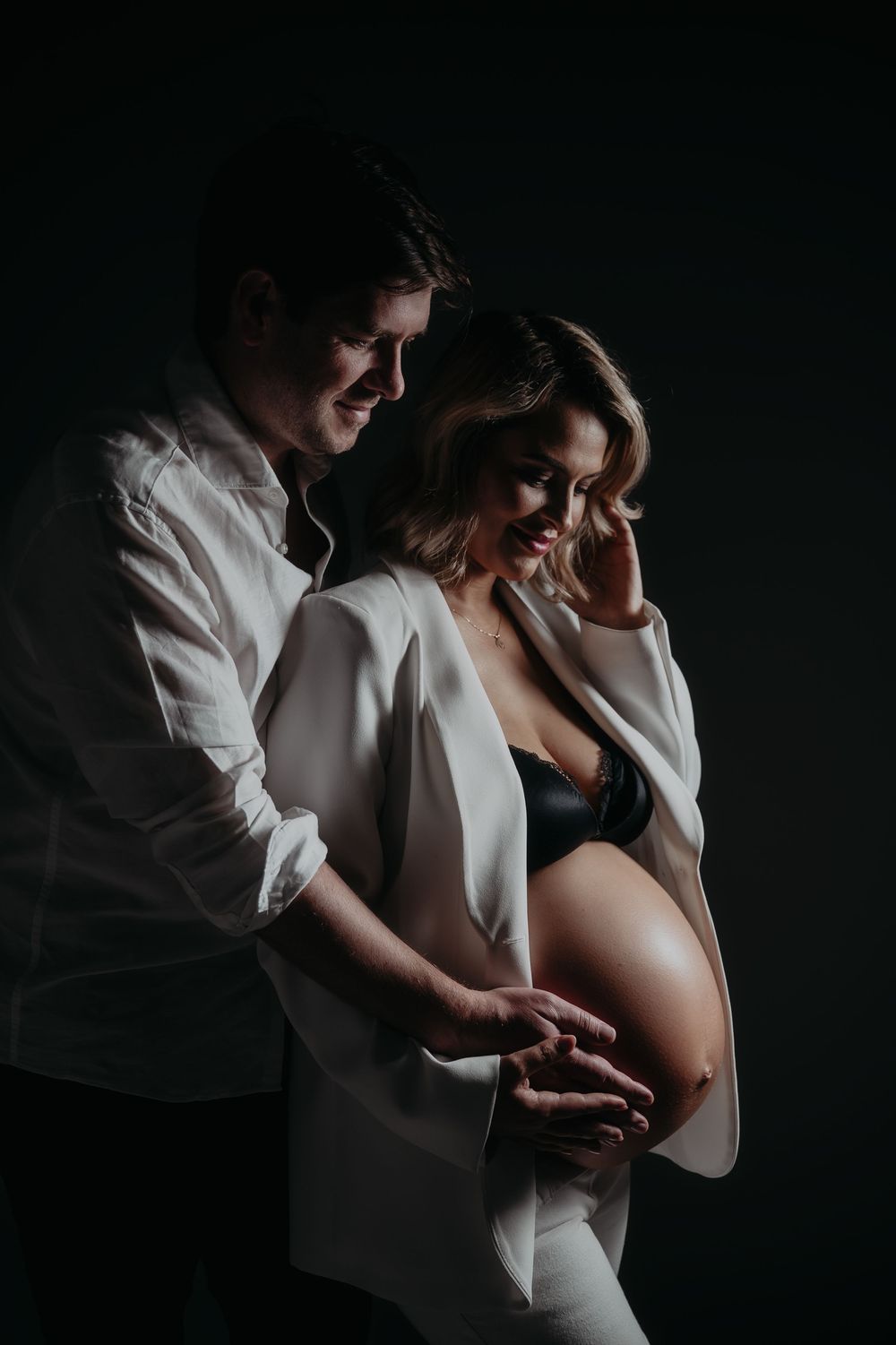 Couple tenderly cradling pregnant belly in dramatic dark studio maternity portrait shoot.