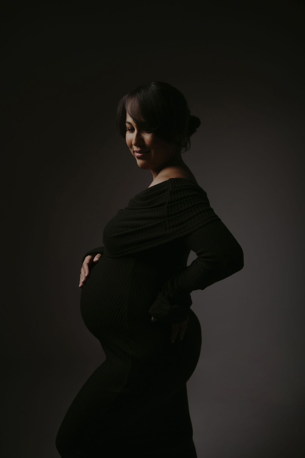 Pregnant woman in black dress cradling her baby bump, smiling in a dramatic dark studio portrait.