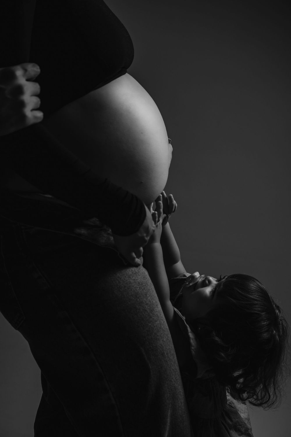Black and white maternity photo of pregnant woman with partner tenderly kissing her baby bump in studio.