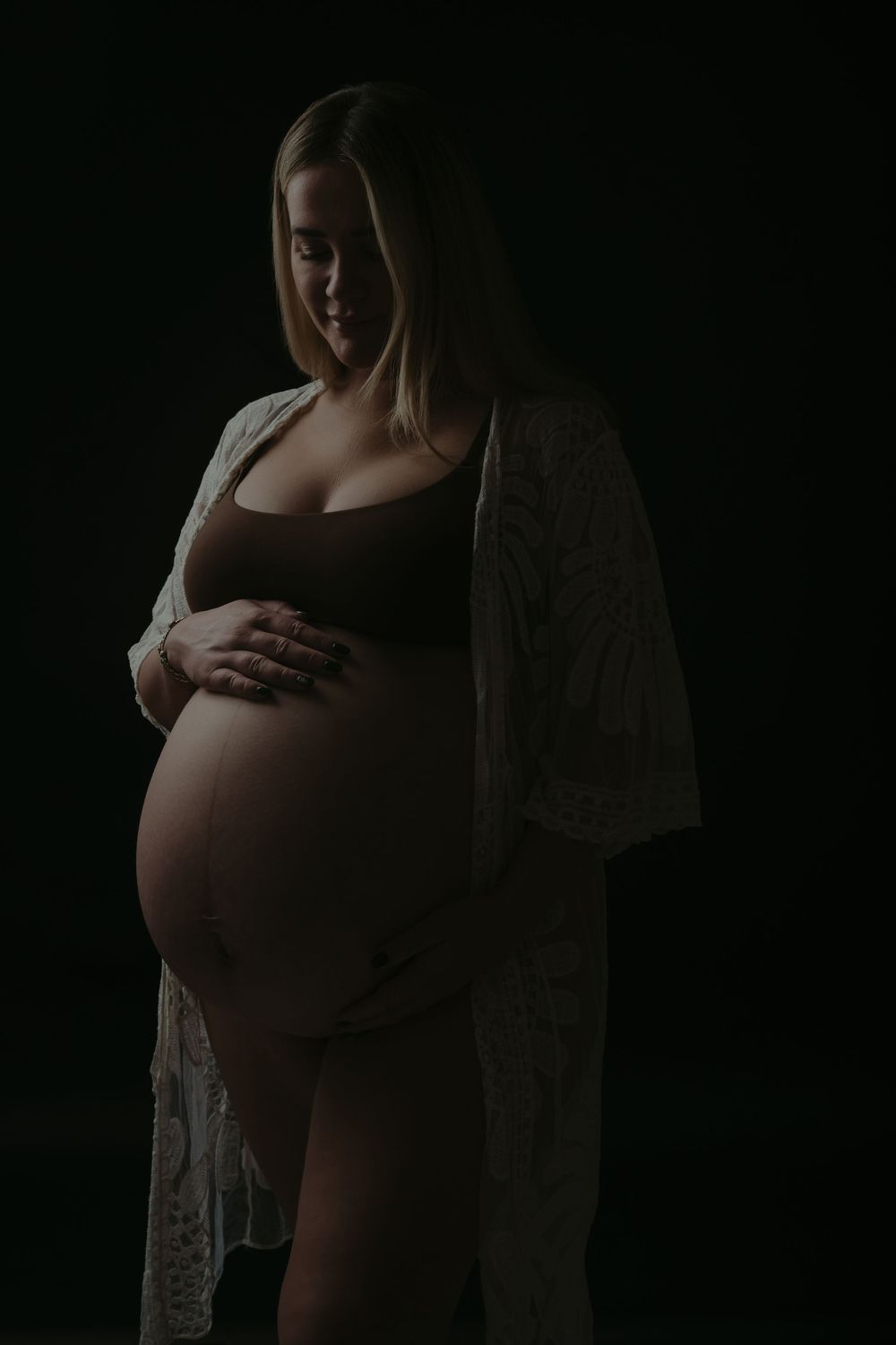 Pregnant woman in dark studio holding baby bump, wearing lace robe and brown bodysuit, maternity photography.