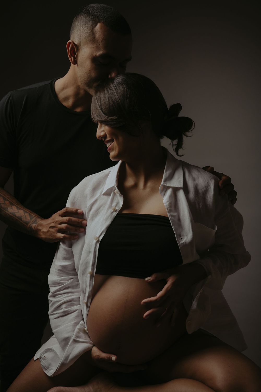 A couple poses tenderly in a dark studio, the pregnant woman cradling her bare belly while her partner embraces her from behind.