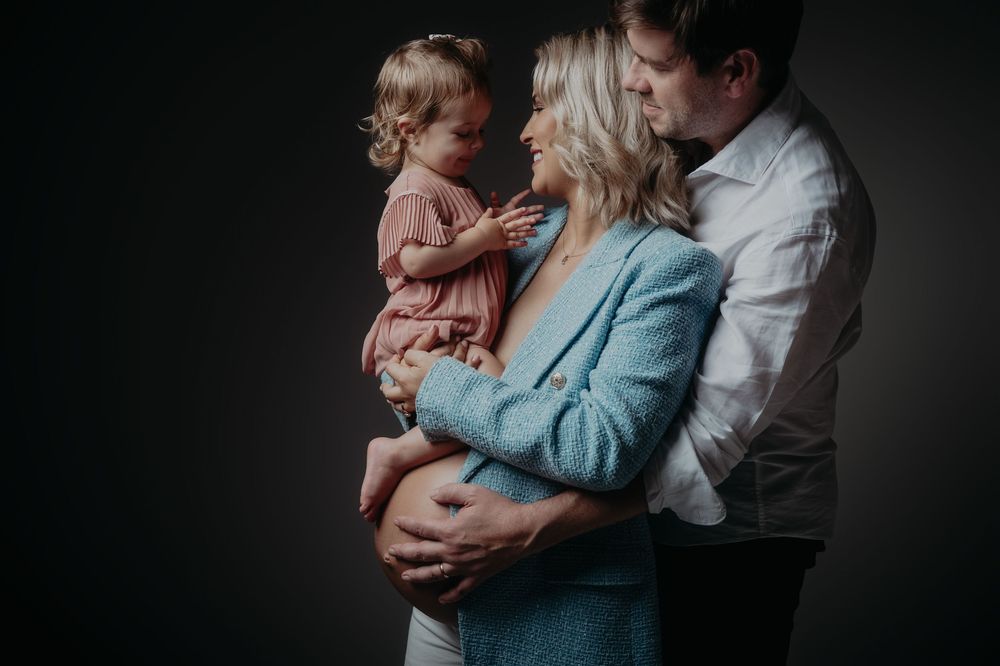 A pregnant woman in a blue blazer holds a toddler girl while a man embraces them both in a studio maternity portrait.