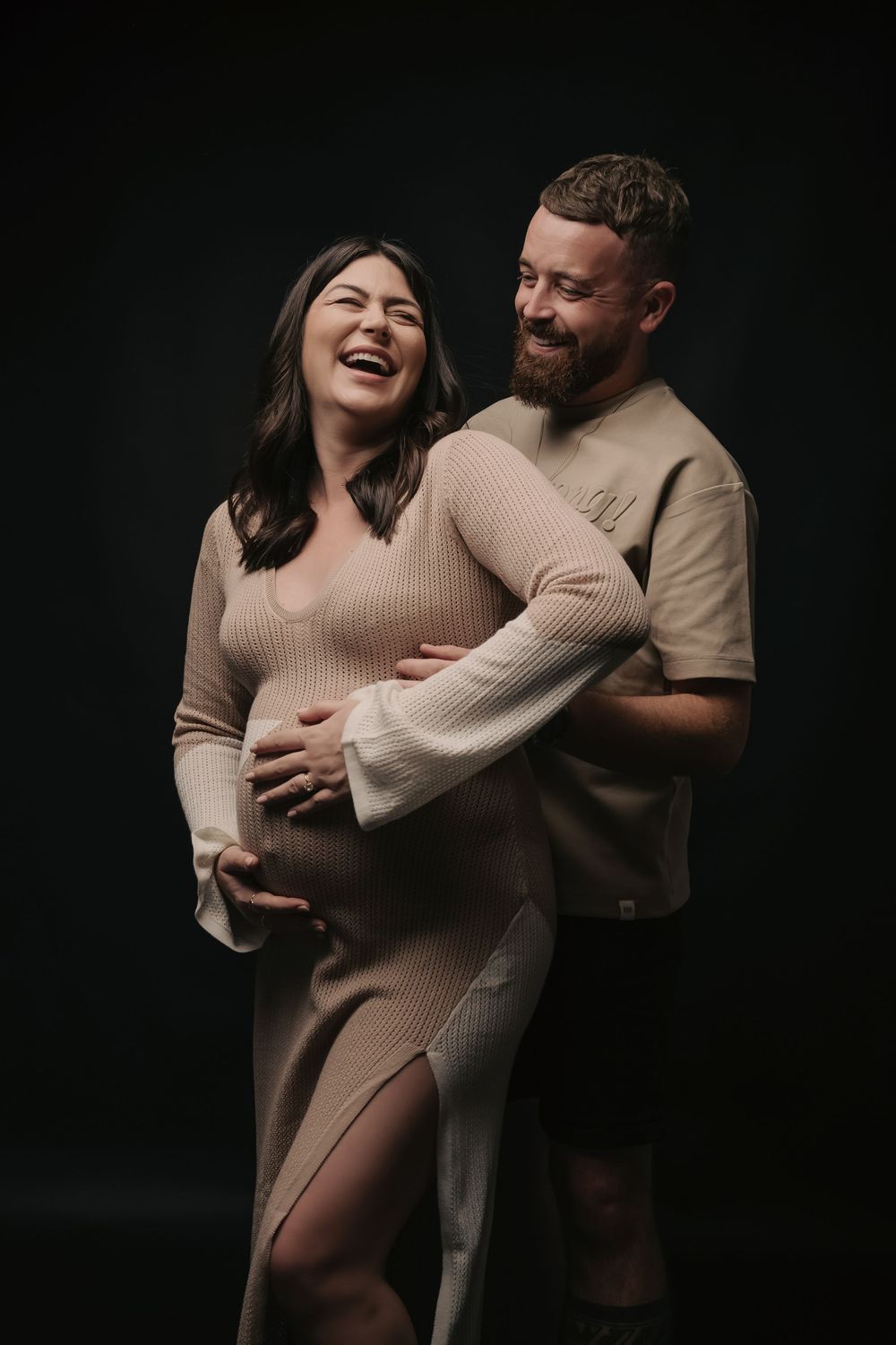 Couple in matching beige outfits laughing during maternity photo shoot on dark background.