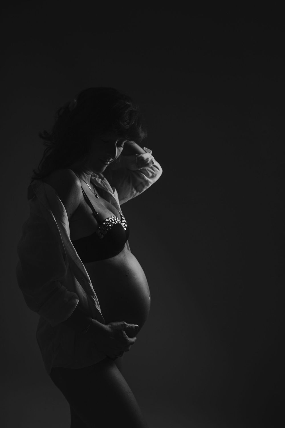 Black and white silhouette of a pregnant woman in lingerie cradling her baby bump against a dark background.