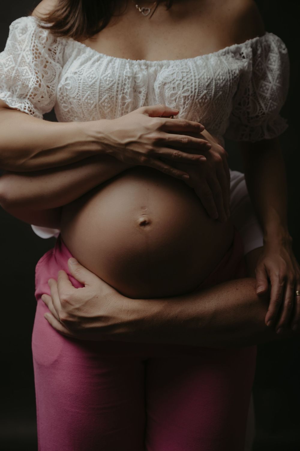 Pregnant woman in white lace top and pink pants cradling baby bump with partner's hands wrapped around her.