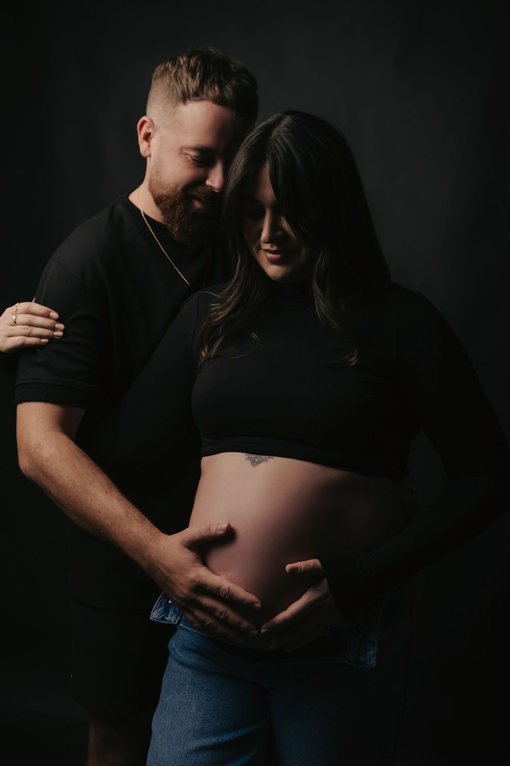 A couple tenderly cradles a pregnant belly in a dark studio, the man embracing the woman from behind in matching black outfits.
