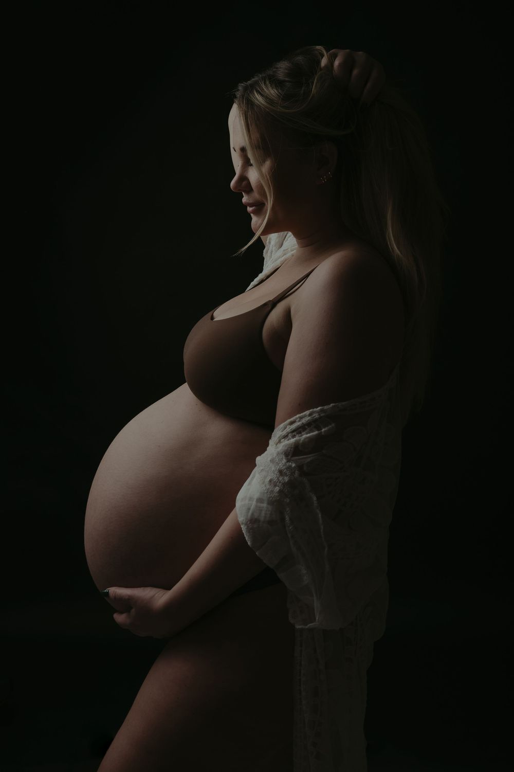Pregnant woman in brown bra holding baby bump, side profile against dark background in artistic maternity photo.
