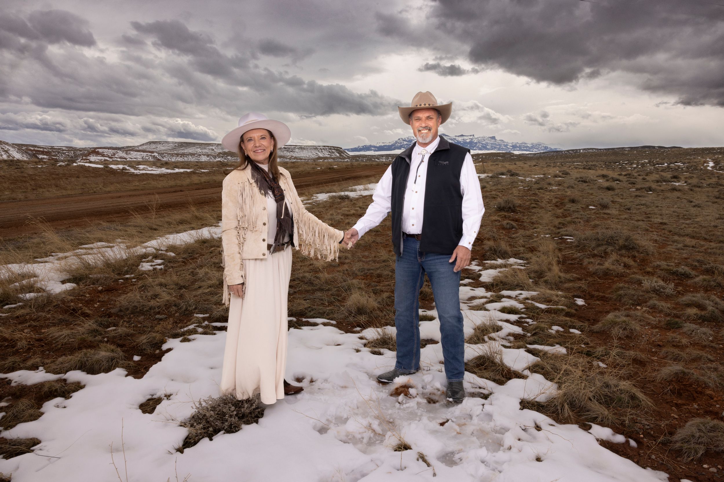 Couple dressed in western attire pose together on snowy ground with moody sky background.