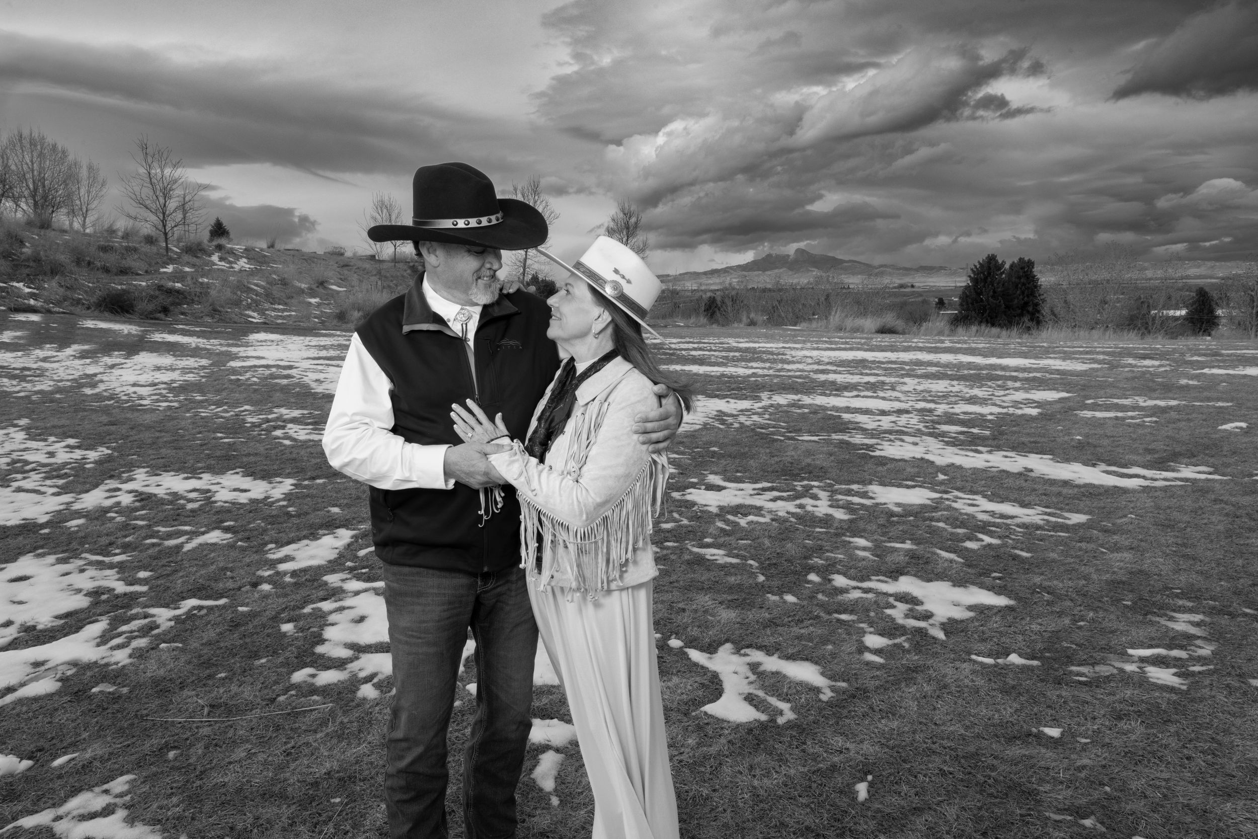 Black and white western-themed couple portrait in snowy field with dramatic clouds.