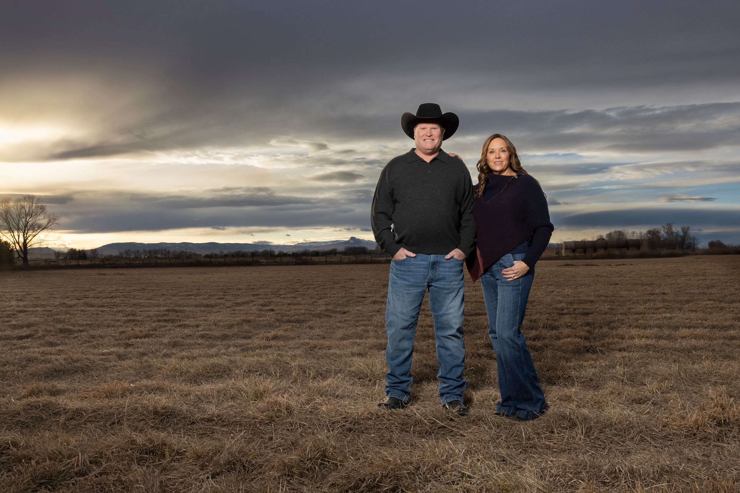 Couple in western wear standing together in field at dusk with dramatic sky.