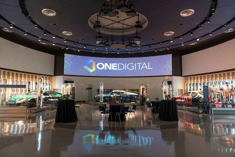 Modern corporate event space with One Digital logo display on large screen and circular lighting fixture overhead.